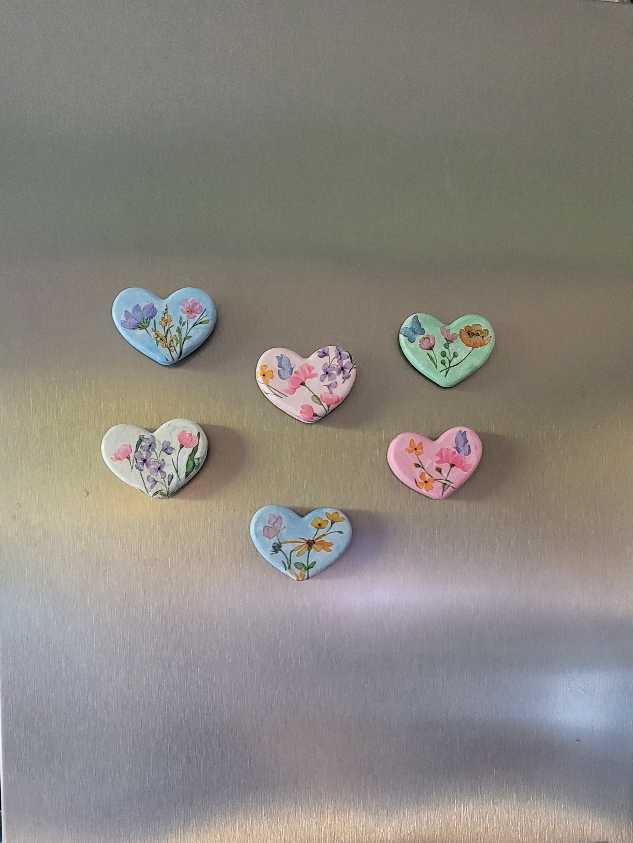 Six heart-shaped floral magnets in pastel colors are displayed on the stainless steel door of a side-by-side refrigerator.