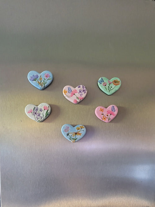 Six heart-shaped floral magnets in pastel colors are displayed on the stainless steel door of a side-by-side refrigerator.