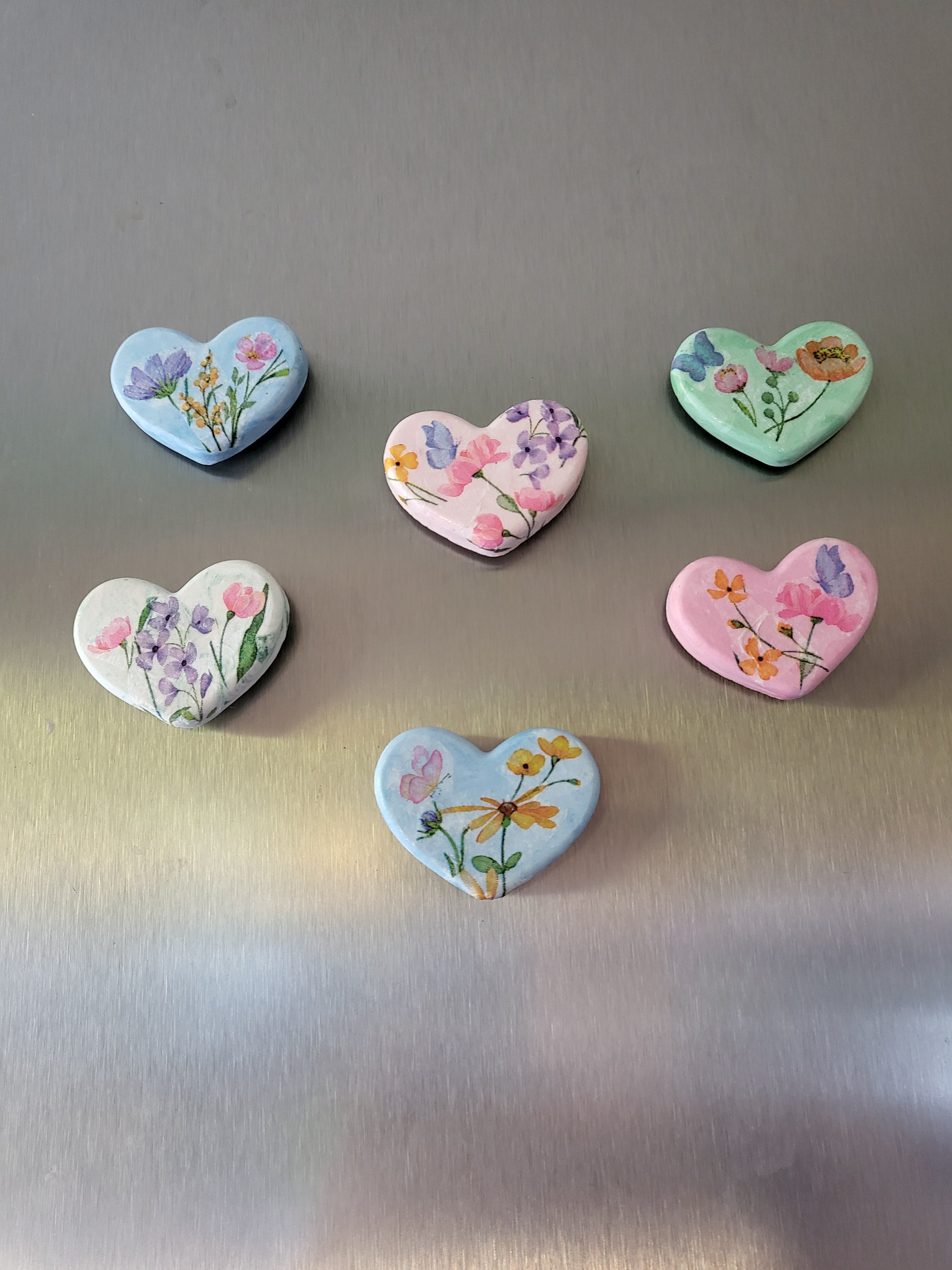 Six heart-shaped refrigerator magnets with colorful floral designs, arranged on a brushed stainless steel surface.